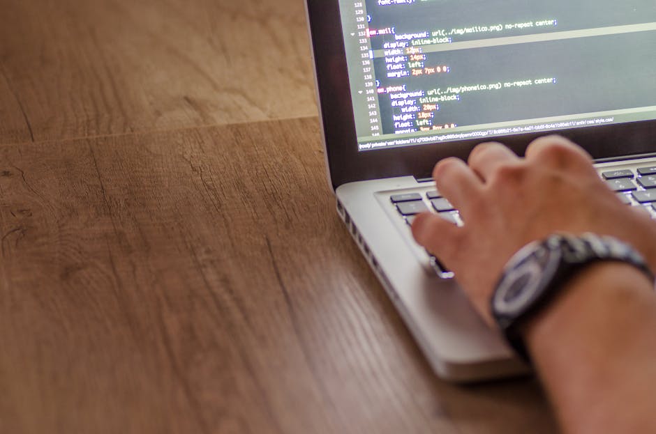 A close-up view of a person's hand typing on a laptop keyboard placed on a wooden surface. The laptop screen displays lines of code with a dark background and colorful syntax highlighting. The individ