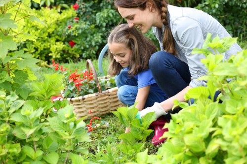 A woman and a young girl are gardening together in a lush, well-maintained garden, surrounded by various green plants, including bushes, hedges, and flowering shrubs. The woman, with long brown hair tied back, is crouched down on the grass, observing the girl who is holding a wicker basket filled with small red flowers. The garden features a neatly kept lawn with dense green grass, bordered by a mix of flowering plants and leafy bushes. In the background, there are mature trees and a variety of foliage, creating a vibrant, natural outdoor space. The scene appears to take place on a bright, clear day, with natural sunlight illuminating the scene. This setting reflects a well-organized, inviting outdoor environment suitable for family gardening activities, natural landscaping, and outdoor maintenance, aligning with professional gardening practices from Gardener Hayes, serving the Hayes area postcodes.