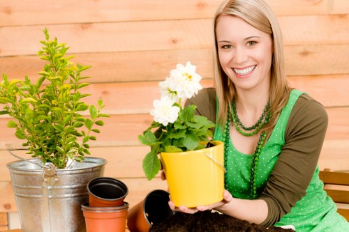 The image shows a young woman with fair skin and shoulder-length blonde hair smiling while holding a small yellow ceramic pot with a flowering plant that has white blooms and green foliage. She is seated outdoors on a wooden patio with a warm, natural wood background, and is dressed in a green polka-dot top with a brown cardigan. In front of her on the patio are several empty flower pots made of terracotta and plastic, indicating gardening activity. To her left, there is a small potted plant with bright green, dense foliage in a metallic container, possibly a shrub or herbal plant. The scene suggests a sunny day with natural outdoor lighting, and the overall setting exudes a welcoming, gardening-friendly atmosphere. This image reflects a gardening practice related to planting, potting, and outdoor garden maintenance, which aligns with services offered by Gardener Hayes in the local area near Hayes or nearby South West London. The composition emphasizes healthy plants, garden tools, and a clean, well-maintained outdoor space suitable for gardening and landscaping activities.