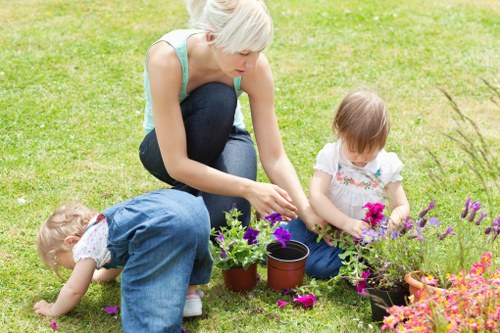 A woman with short blonde hair, wearing a light blue sleeveless top and dark blue jeans, is kneeling on a lush, well-maintained green lawn in a garden. She is engaged in gardening activities, tending to potted plants with vibrant purple, pink, and orange flowers, which are arranged in black pots on the grass. Two young children, a boy and a girl, are also present; the boy, dressed in a blue denim shirt and light-colored trousers, is bent over, closely examining or helping with a plant, while the girl, wearing a white dress with pink floral embroidery, sits nearby watching attentively. The garden features a tidy and organized layout with a variety of flowering plants, contributing to a colourful and natural outdoor space. The background shows part of the garden with more plants and greenery, under a bright and partly cloudy sky, suggesting a pleasant weather day suitable for outdoor gardening. This scene exemplifies typical gardening activities that Gardener Hayes might support, highlighting the importance of a well-kept lawn and vibrant, flourishing flower beds in a residential garden setting near Hayes.